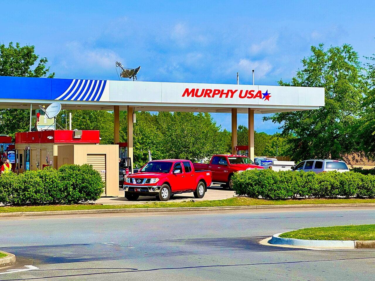 A Murphy USA gas station with several cars parked at the pumps, including a red pickup truck in front. Trees and bushes surround the station on a sunny day.