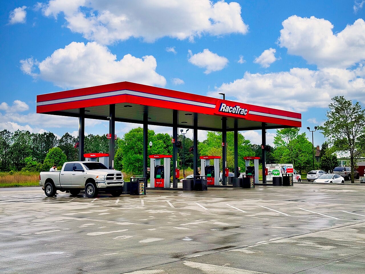 A white pickup truck is parked at a RaceTrac gas station with multiple red and black fuel pumps under a red canopy on a clear, sunny day with scattered clouds. Trees and greenery surround the station.