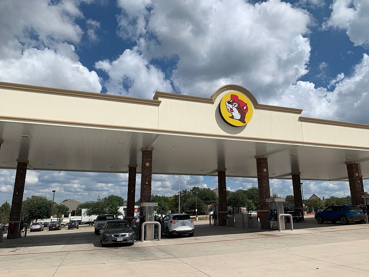 Cars are parked at gas pumps under a large Buc-ee’s gas station canopy, featuring the Buc-ee’s beaver logo, on a partly cloudy day with trees and houses in the background.