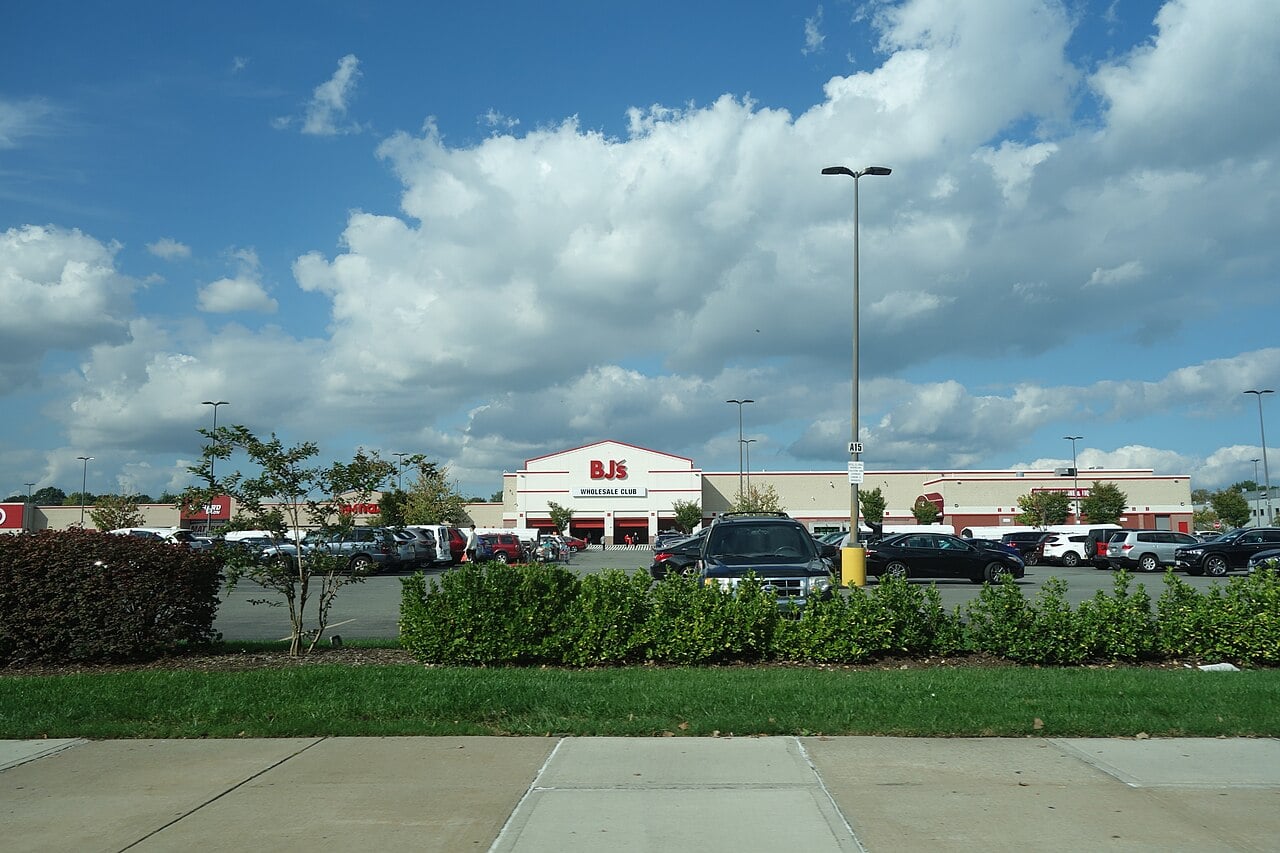 A BJ’s Wholesale Club store with a large parking lot filled with cars, green shrubs, and a partly cloudy blue sky overhead.