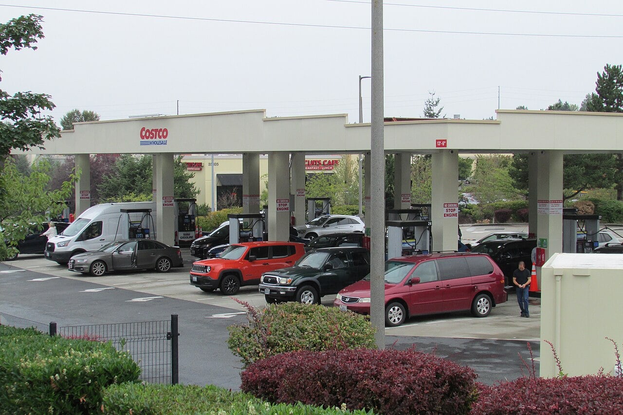 Several vehicles, including cars, SUVs, and a van, are parked at gas pumps under a Costco Gasoline station canopy on a cloudy day. People are refueling their vehicles, and greenery surrounds the station.