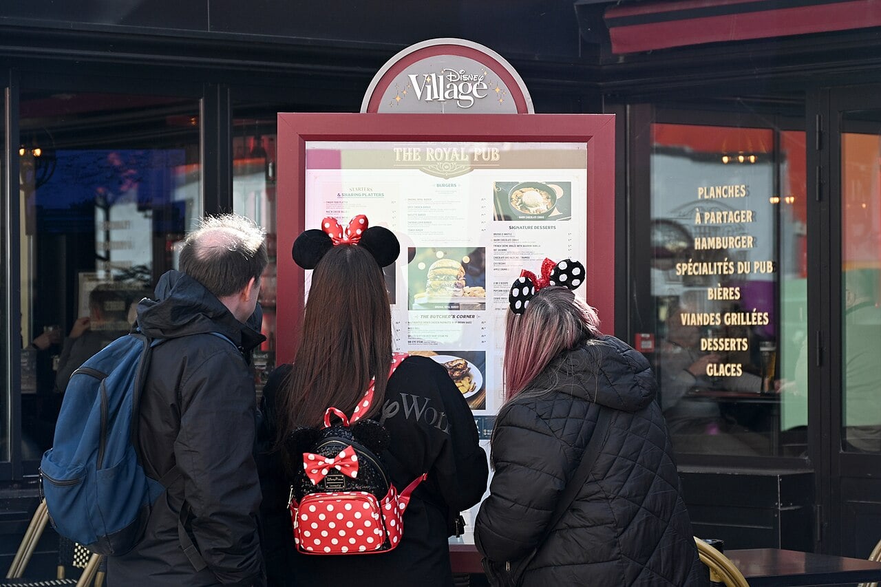 Three people wearing jackets and Disney-themed mouse ear headbands look at a menu displayed outside The Royal Pub at Disney Village. The menu lists various food options in both English and French.