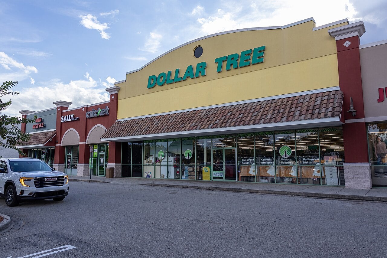 A shopping plaza with a large Dollar Tree store in the center, featuring green signage and glass windows. There are adjacent stores, a parked SUV, and a mostly empty parking lot under a partly cloudy sky.