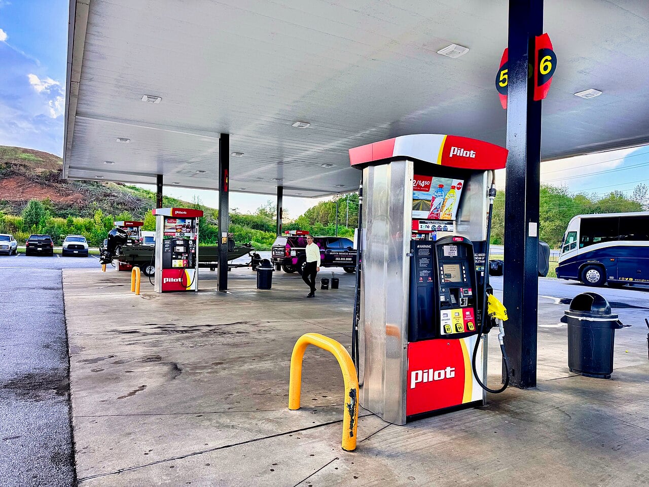 A Pilot gas station with multiple fuel pumps, cars parked at the pumps, and a person walking across the forecourt. A hill and trees are in the background, with a cloudy sky above.