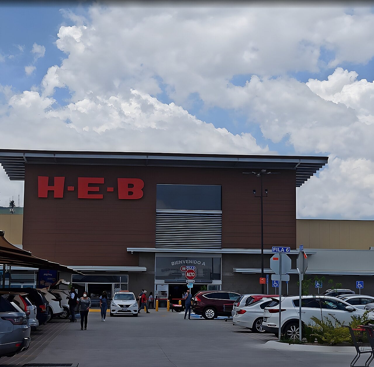 The image shows the front entrance of an H-E-B grocery store with several parked cars and people walking toward the entrance under a partly cloudy sky.