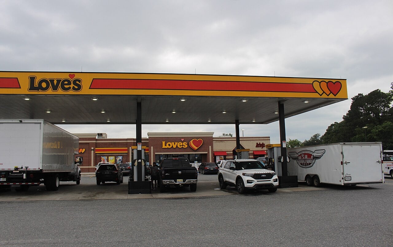 Several vehicles, including trucks and SUVs, are parked under a yellow and red Love’s gas station canopy, with the convenience store visible in the background on an overcast day.
