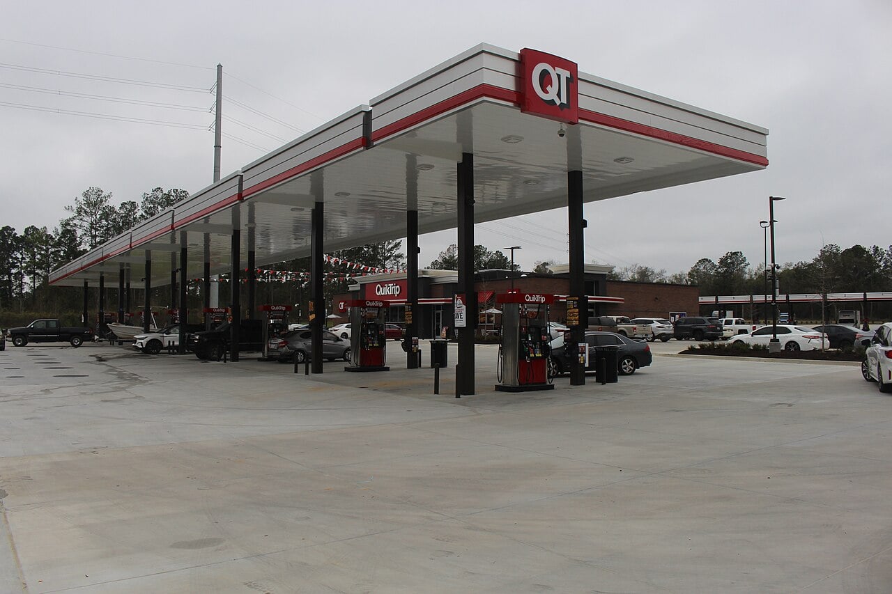 A QuikTrip gas station with multiple fuel pumps under a large canopy, several cars parked around, and a convenience store building in the background on a cloudy day.