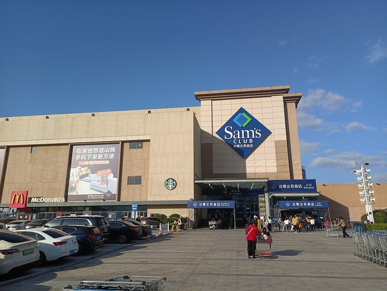 A large Sam's Club store with Chinese signage is shown under a blue sky. The building also has McDonald's and Starbucks logos. Several cars are parked in front, and people are walking near shopping carts.