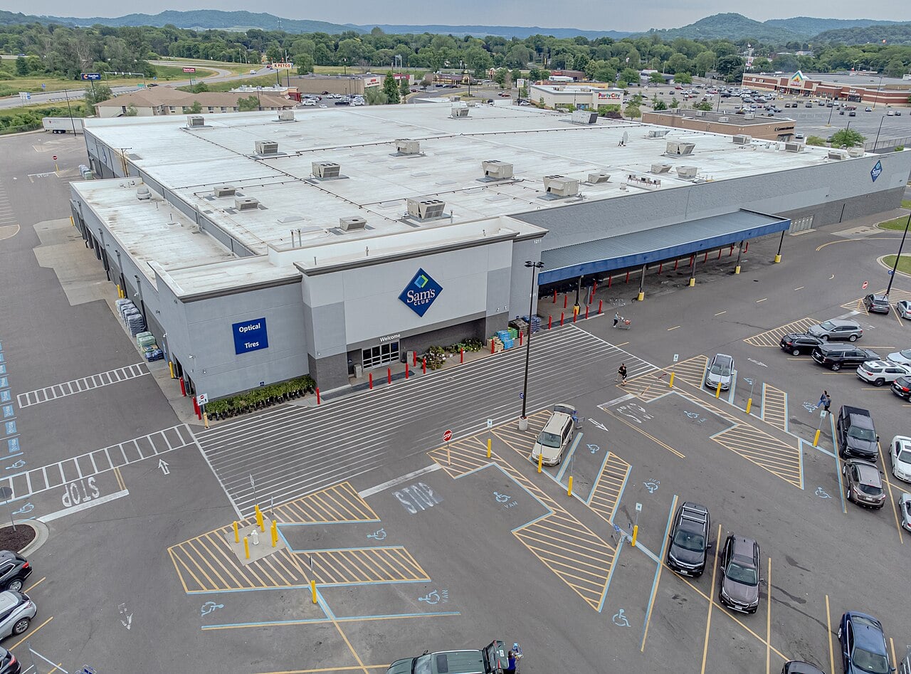 Aerial view of a large Sam's Club store with a busy parking lot, multiple cars parked, and mountains and other businesses visible in the background. The store entrance is visible with shoppers nearby.