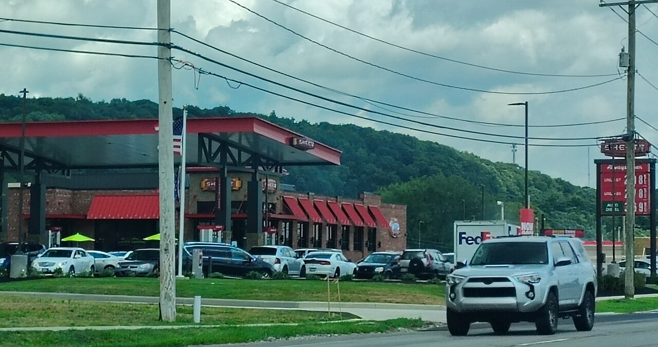 A busy gas station with a red canopy and a convenience store is surrounded by parked cars. A white SUV drives by in the foreground, and there are green hills and cloudy skies in the background.