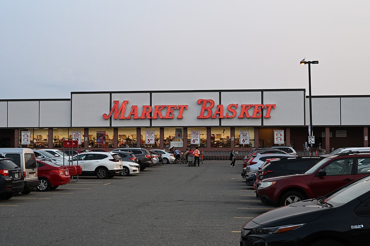 A parking lot filled with cars in front of a Market Basket grocery store at dusk, with the store’s bright red sign and large windows visible.