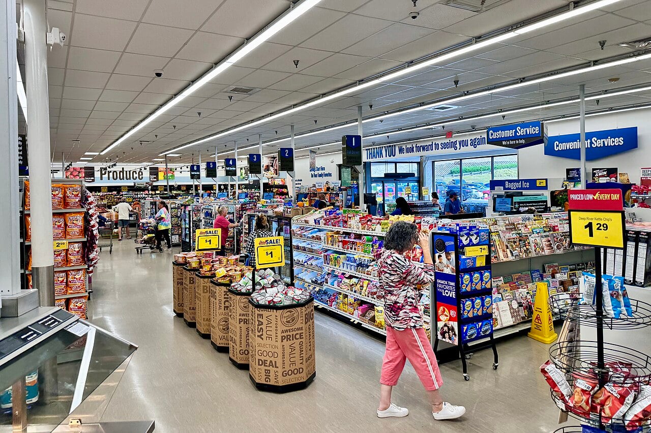 A woman walks through a brightly lit grocery store with displays of snacks, magazines, and produce. Signs hang above aisles, and other shoppers are seen in the background near checkout counters.