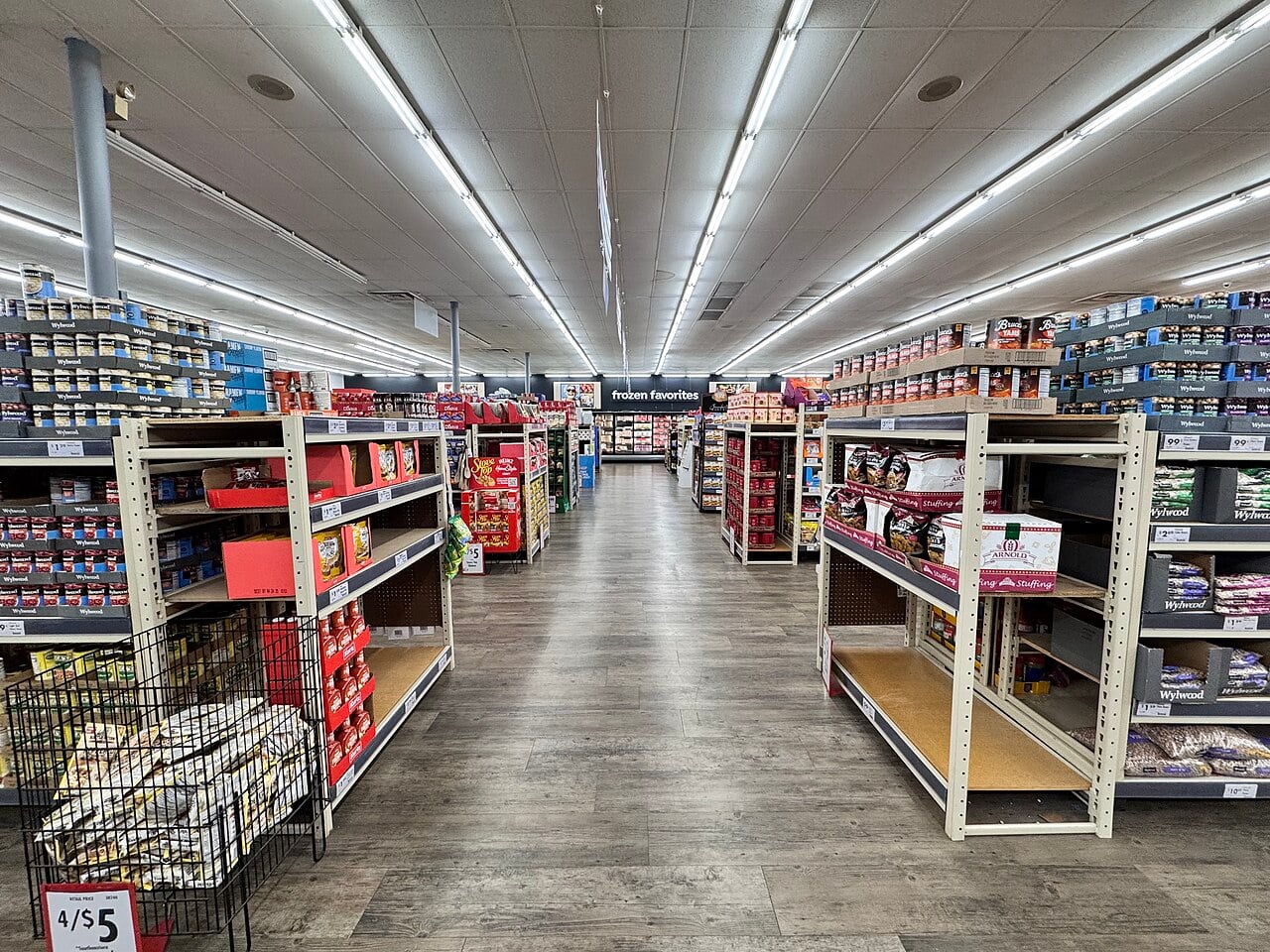 A grocery store aisle with shelves stocked with various food products on both sides; some shelves are partially empty. The floor is wood-patterned, and overhead fluorescent lights brighten the store.