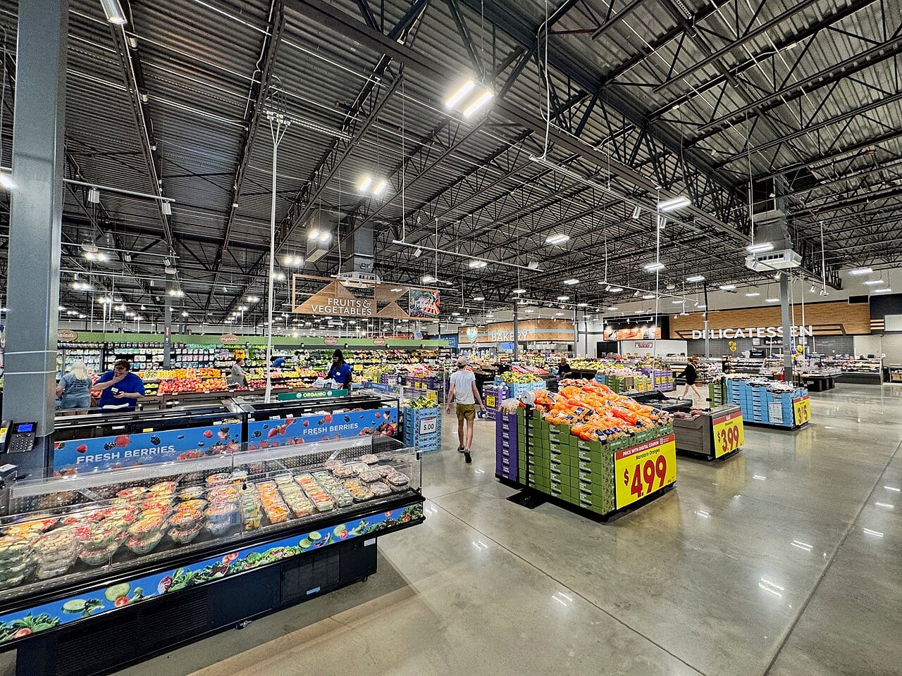 A spacious supermarket produce section with fresh fruits and vegetables on display, shoppers browsing, and signs for berries, fruit, and the delicatessen; prices and bright lighting are visible.