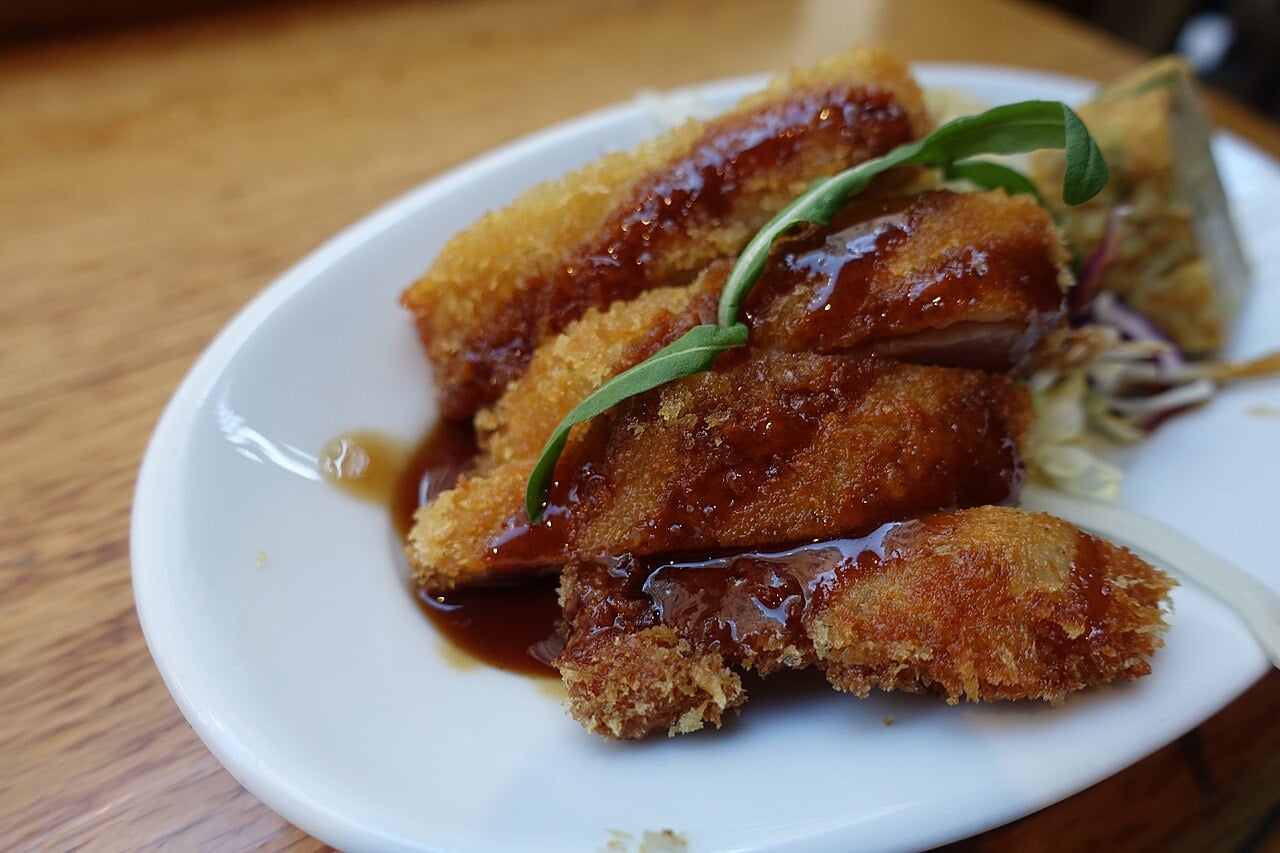 A close-up of breaded and fried slices of meat drizzled with brown sauce, served on a white plate with coleslaw and garnished with a green sprout.