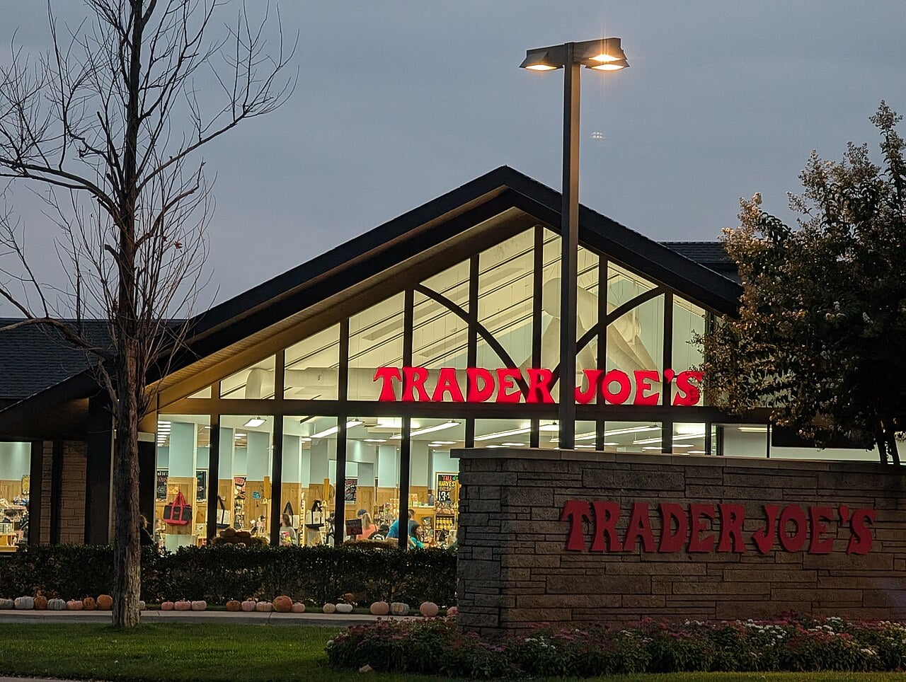 A Trader Joe's grocery store at dusk, with large windows revealing a brightly lit interior. The red Trader Joe's sign is illuminated on the building and on a stone sign in front. Trees and a lamp post are visible outside.