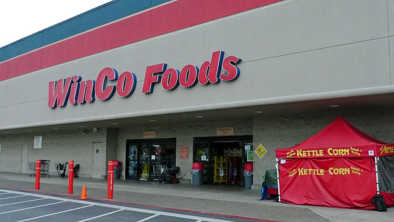 WinCo Foods storefront with automatic glass doors, shopping carts near the entrance, and a red tent labeled "Kettle Corn" set up outside on the right. A parking lot and traffic cones are visible in front.