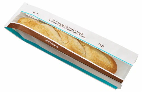 A freshly baked baguette in a white and blue paper bag, partially open to show the golden crust. The packaging has brown and turquoise accents and printed text on top.