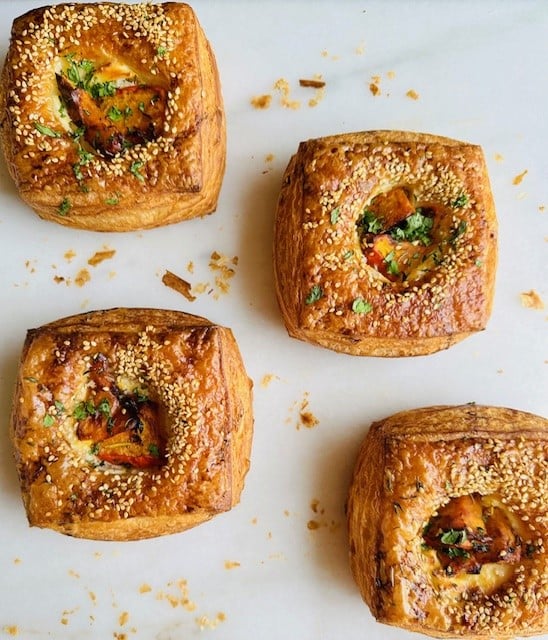 Four square puff pastries topped with sesame seeds and herbs are arranged on a white surface, with a few crumbs scattered around them. The pastries are golden brown and each has a visible filling.