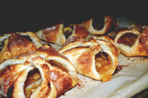 Golden-brown puff pastries with visible apple filling are arranged on parchment paper, with cinnamon sprinkled around them. The pastries look freshly baked and flaky.