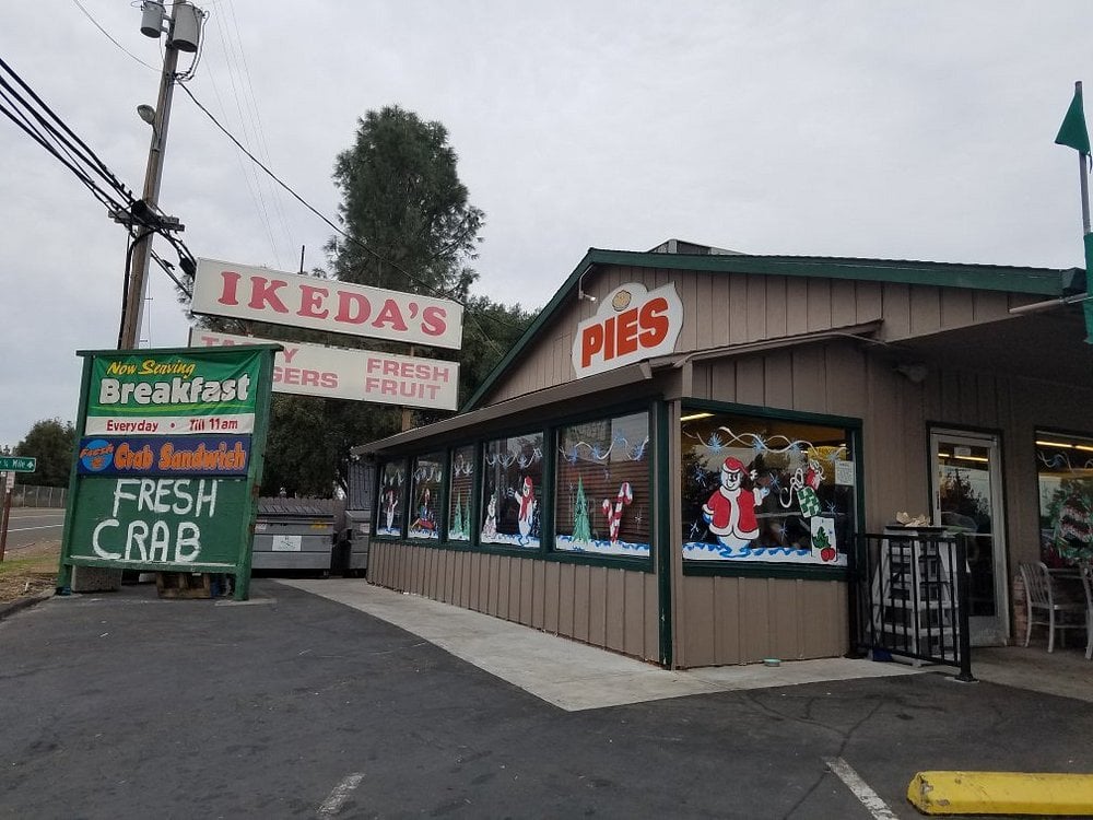 A roadside store called Ikeda's, perfect for road trip food stops, with signs advertising pies, fresh fruit, crab, breakfast, and sandwiches. The windows are decorated with Christmas art and the building sits beside a road under a cloudy sky.