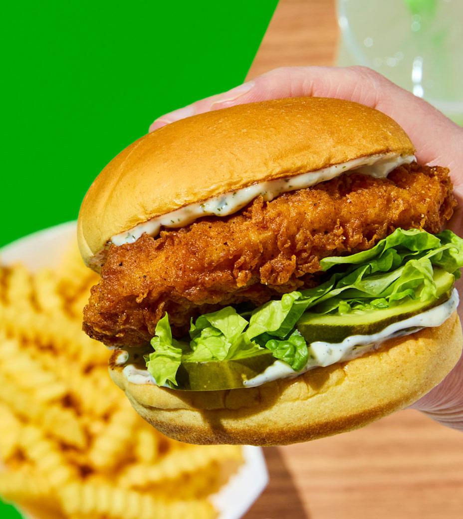 A hand holds a crispy Chicken Sandwich with lettuce and sauce in a soft bun. In the background, crinkle-cut fries and a drink sit on a wooden table against a bright green backdrop.
