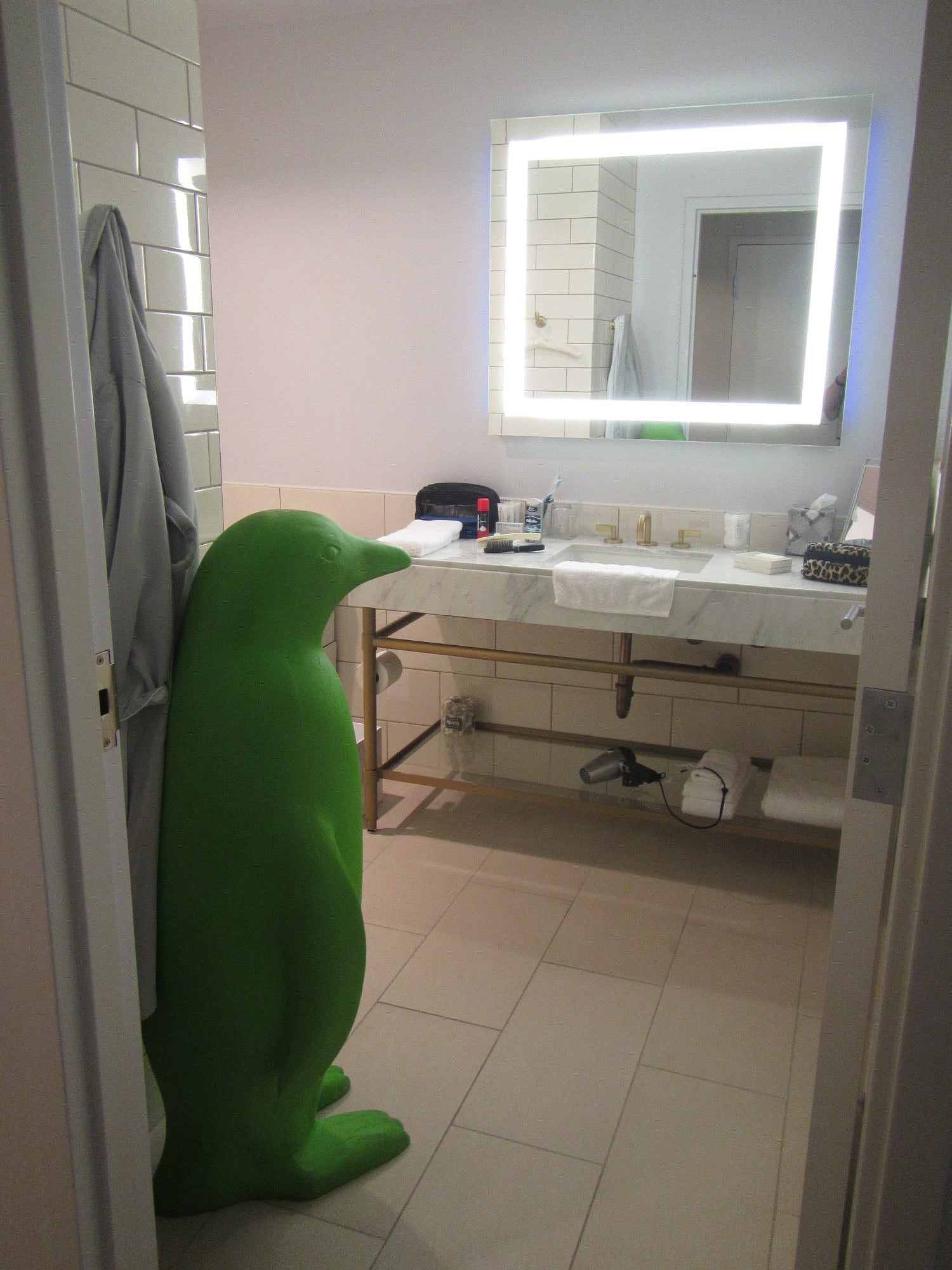 A green, life-size penguin sculpture stands in a modern bathroom near a sink and mirror with bright lights, evoking the quirky charm often found in Small-Town Luxury Hotels, surrounded by toiletries and a grey robe hanging on the wall.