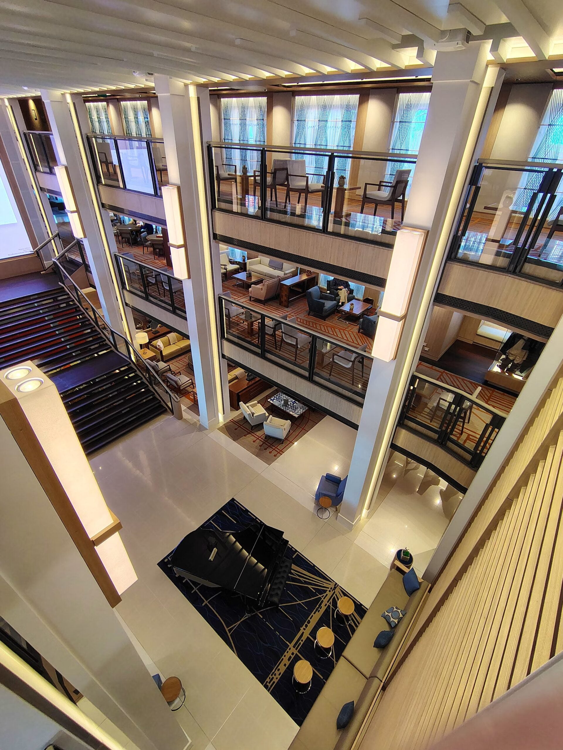 A modern, spacious hotel lobby with multiple floors, open walkways, seating areas, and a black grand piano on a blue carpet in the center—evoking the elegance of a Best Cruise Line 2026 experience. Large windows fill the interior with natural light.