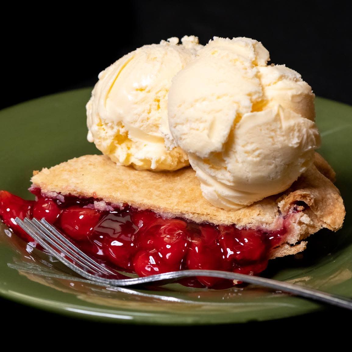 A slice of cherry pie with a golden crust is topped with two scoops of vanilla ice cream on a green plate, with a fork resting in front of the dessert.
