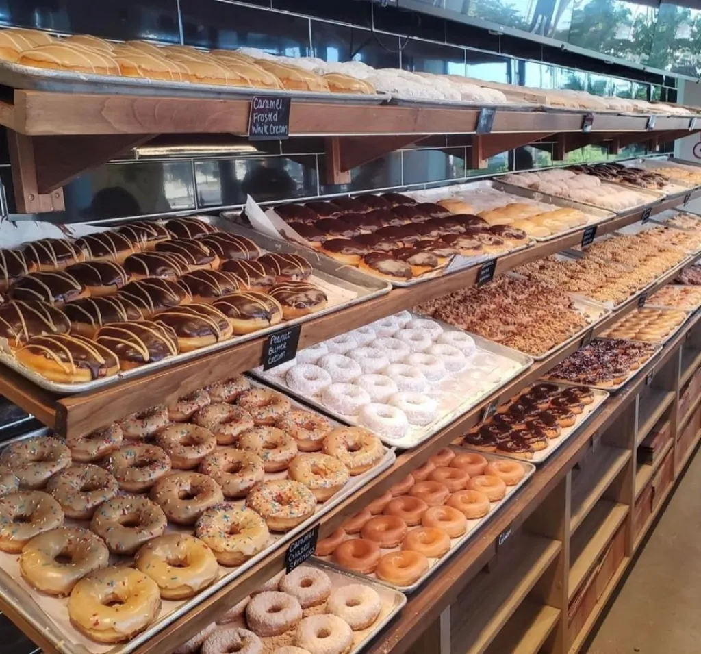 A bakery display filled with trays of various donuts, including chocolate-glazed, sprinkled, powdered sugar, and plain glazed, arranged neatly on shelves.