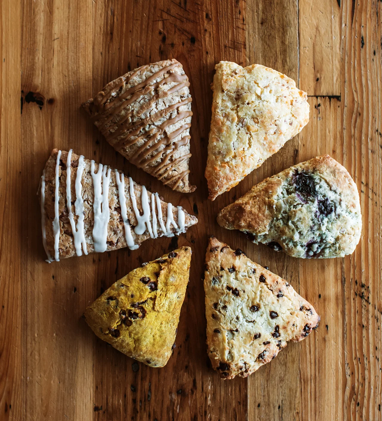 Six assorted scones arranged in a circular pattern on a rustic wooden table, each with different toppings and mix-ins such as glaze, chocolate chips, and fruit.