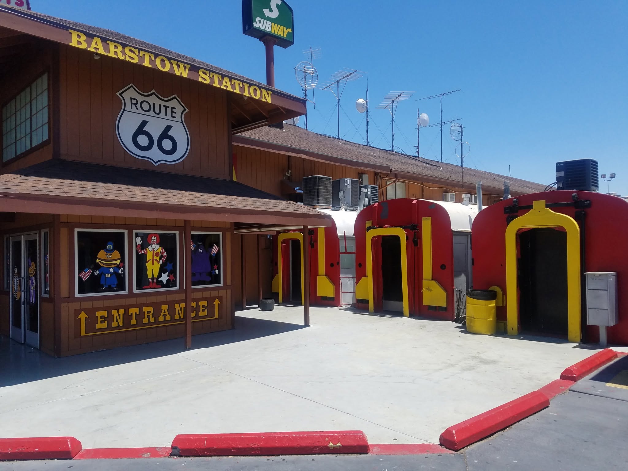 Barstow Station on Route 66, featured among the Coolest McDonald's Locations, showcases a Subway sign, cartoon window art, and red-and-yellow train car structures on a sunny day. A large "Entrance" sign sits above the door.