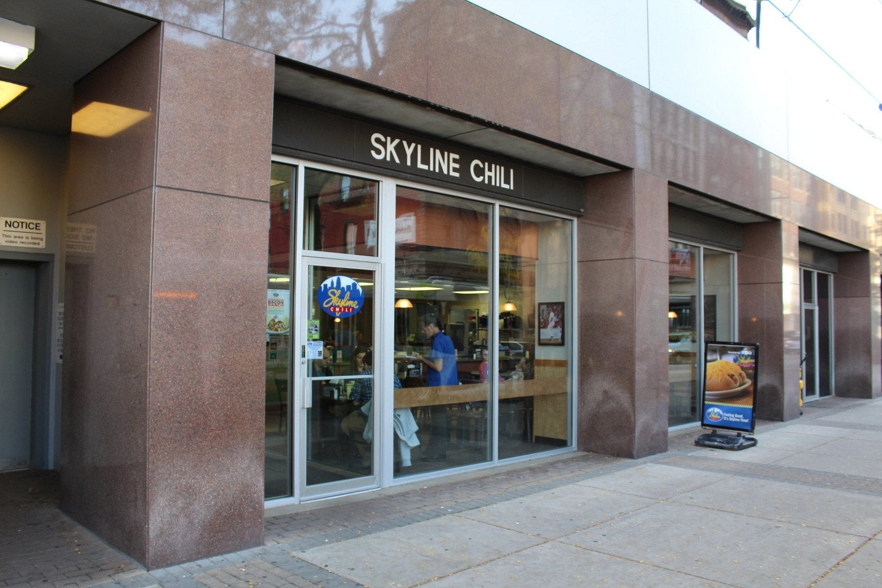The exterior of a Skyline Chili restaurant with large glass windows, a sign above the entrance, and a menu board on the sidewalk—making it a classic spot among memorable road trip food stops. A person is visible inside near the counter.