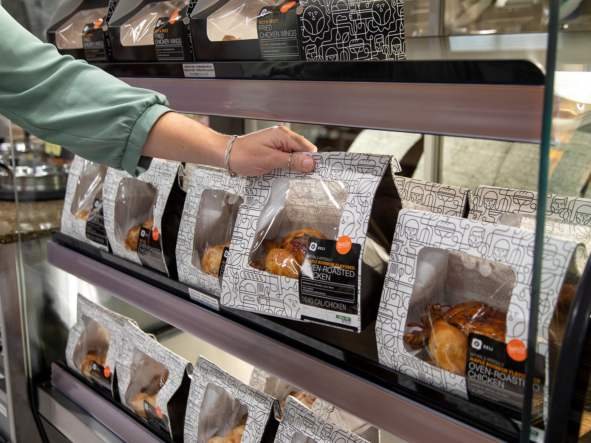 A person’s hand reaches for a packaged rotisserie chicken from a display shelf filled with similar oven-roasted chickens in sealed containers at a grocery store or deli.