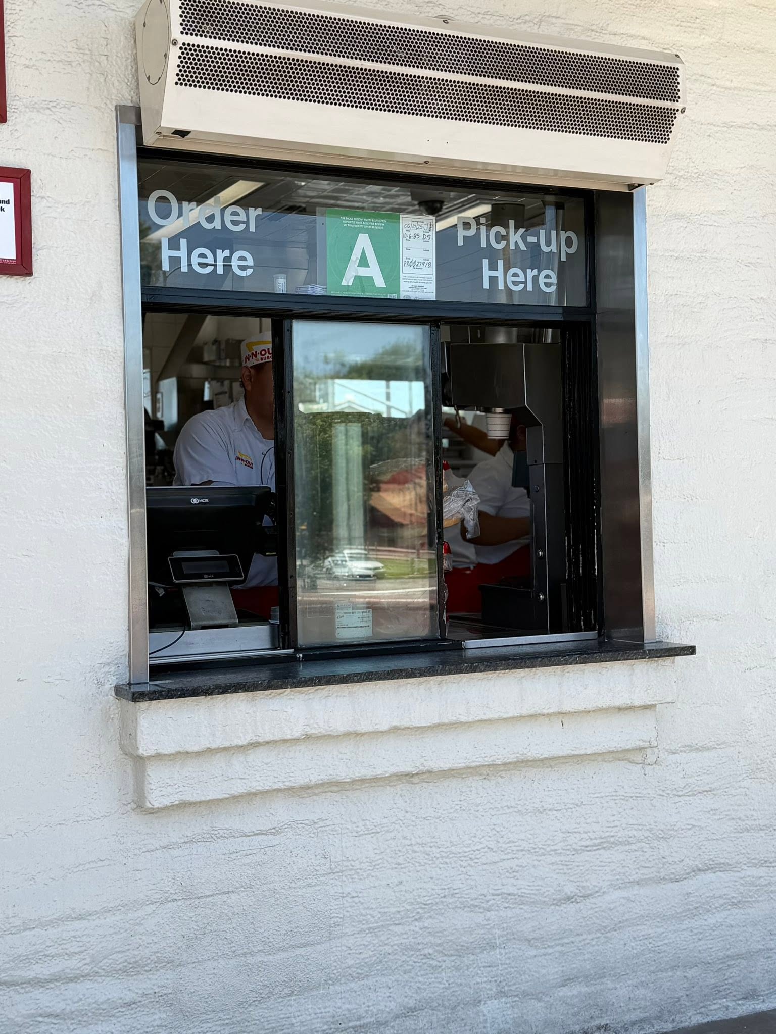 Drive-thru window with "Order Here" and "Pick-up Here" signs above. An employee in uniform is partially visible inside, with equipment and reflections in the glass. White exterior wall and AC unit above the window.