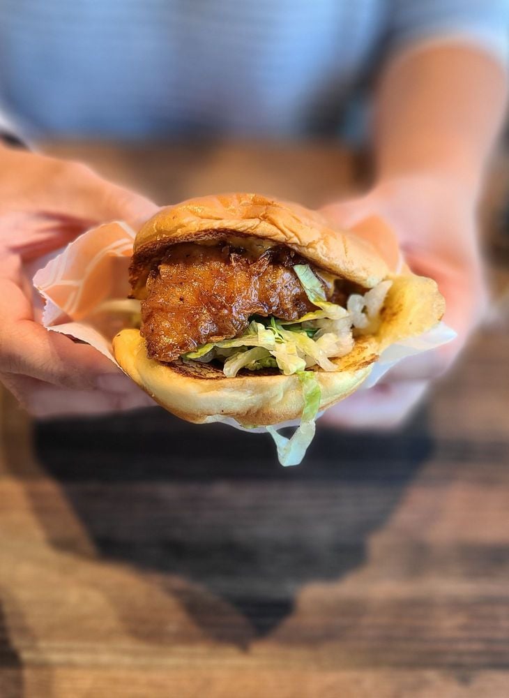 A person holds a crispy chicken sandwich with lettuce and sauce in a soft bun, wrapped partially in paper, over a wooden table.