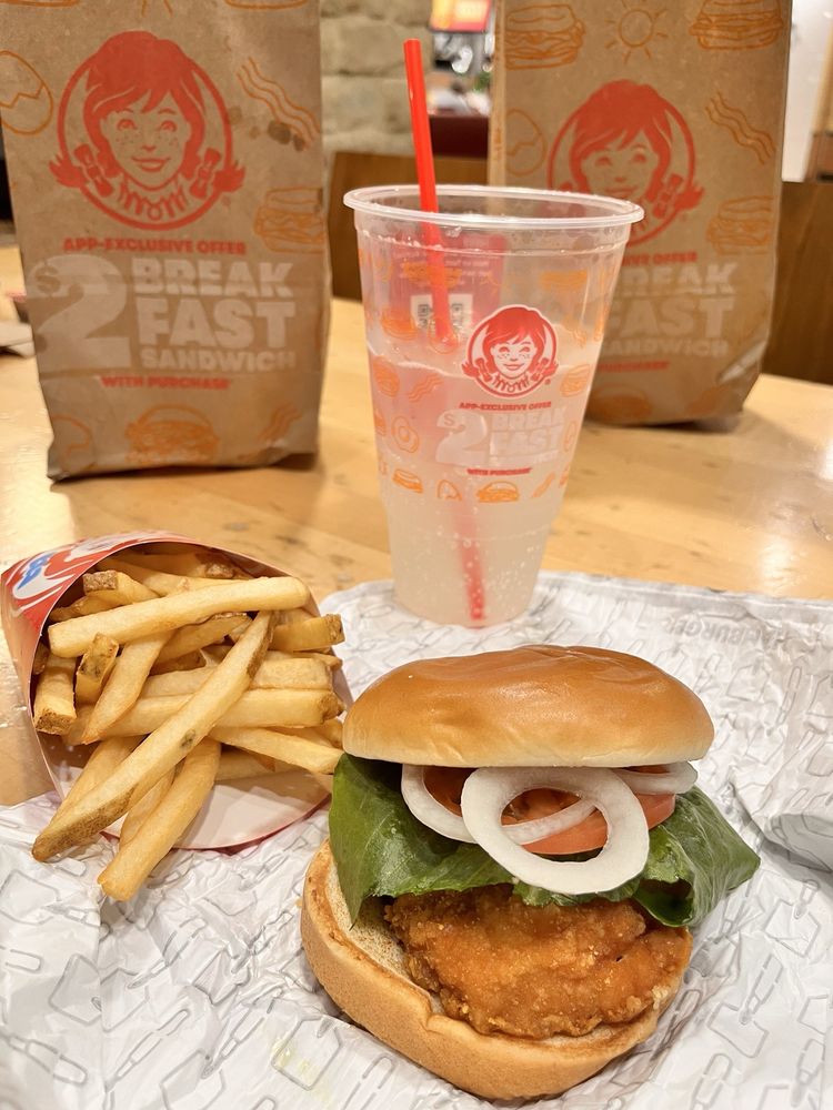A Wendy’s meal on a table featuring a classic Chicken Sandwich with lettuce, tomato, and onion, a serving of fries, a clear cup with a red straw, and two Wendy’s paper bags in the background.