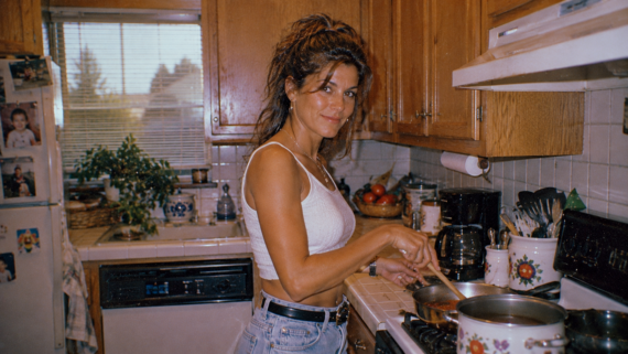 A woman in a white crop top and jeans stands in a cozy kitchen, smiling while stirring meals middle-class moms cooked on the stove. Wooden cabinets, plants, and a sunlit window add warmth to the inviting space.