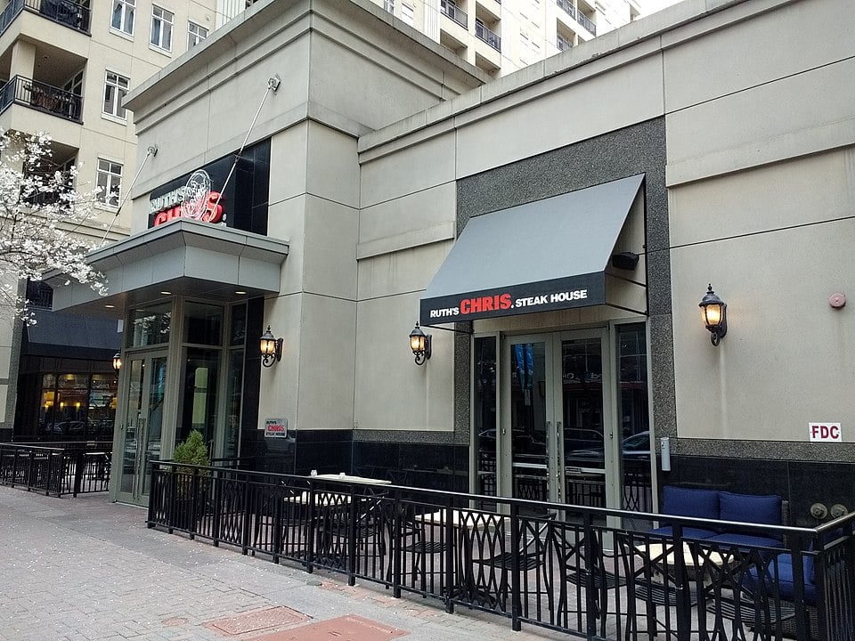 Outdoor view of a Ruth's Chris Steak House with patio seating, black metal fence, and beige building facade. The restaurant's red and white signs are visible above doors and windows.