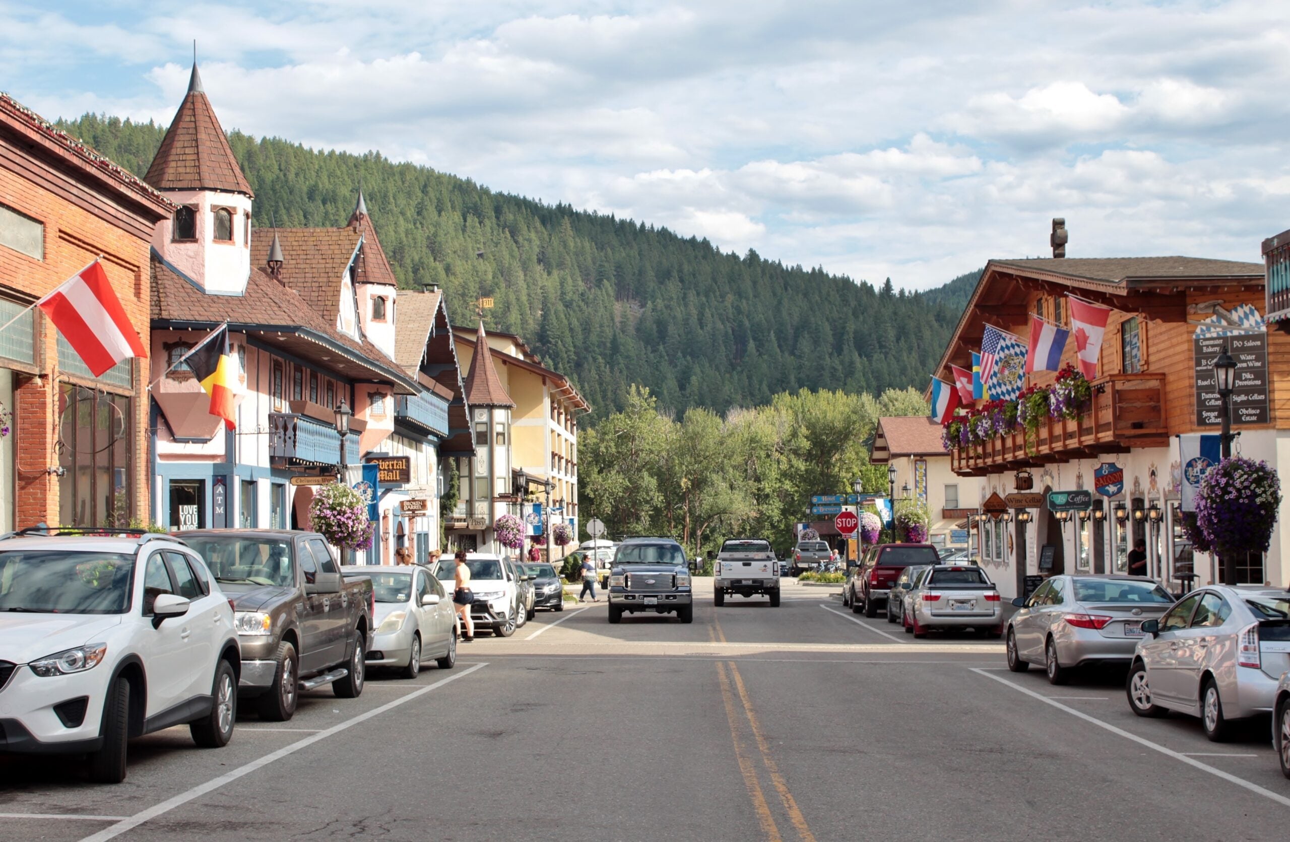 A street scene in a small mountain town with Bavarian-style buildings, colorful flags, parked cars, flower baskets, and forested hills in the background under a partly cloudy sky.