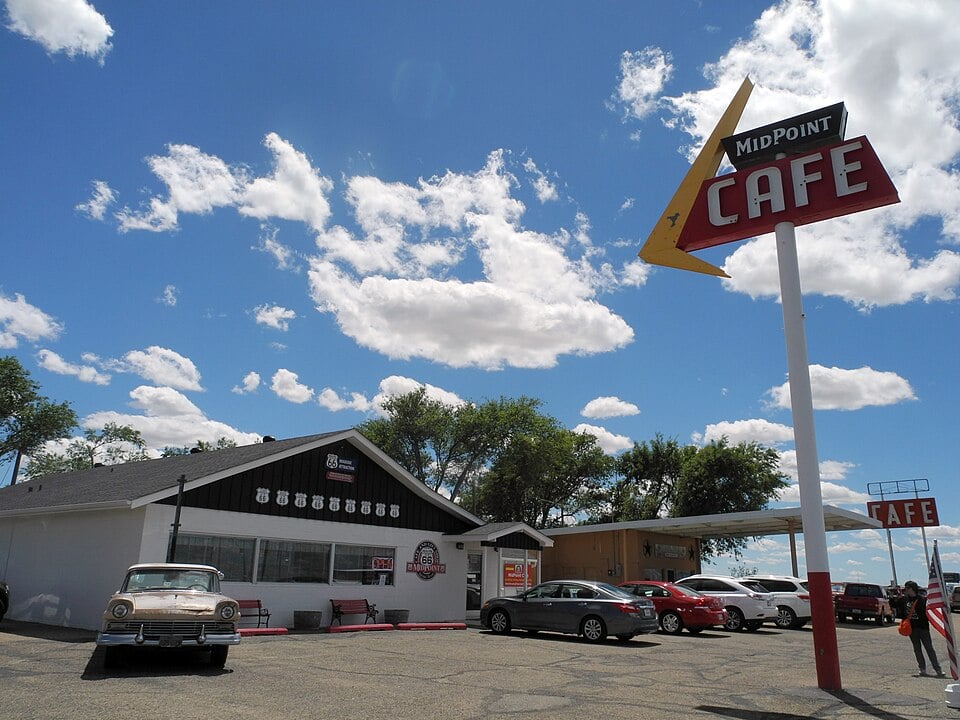 A vintage car is parked outside the Midpoint Cafe, a classic diner with a large retro sign, under a blue sky with scattered clouds. Several other cars are also parked near the building.