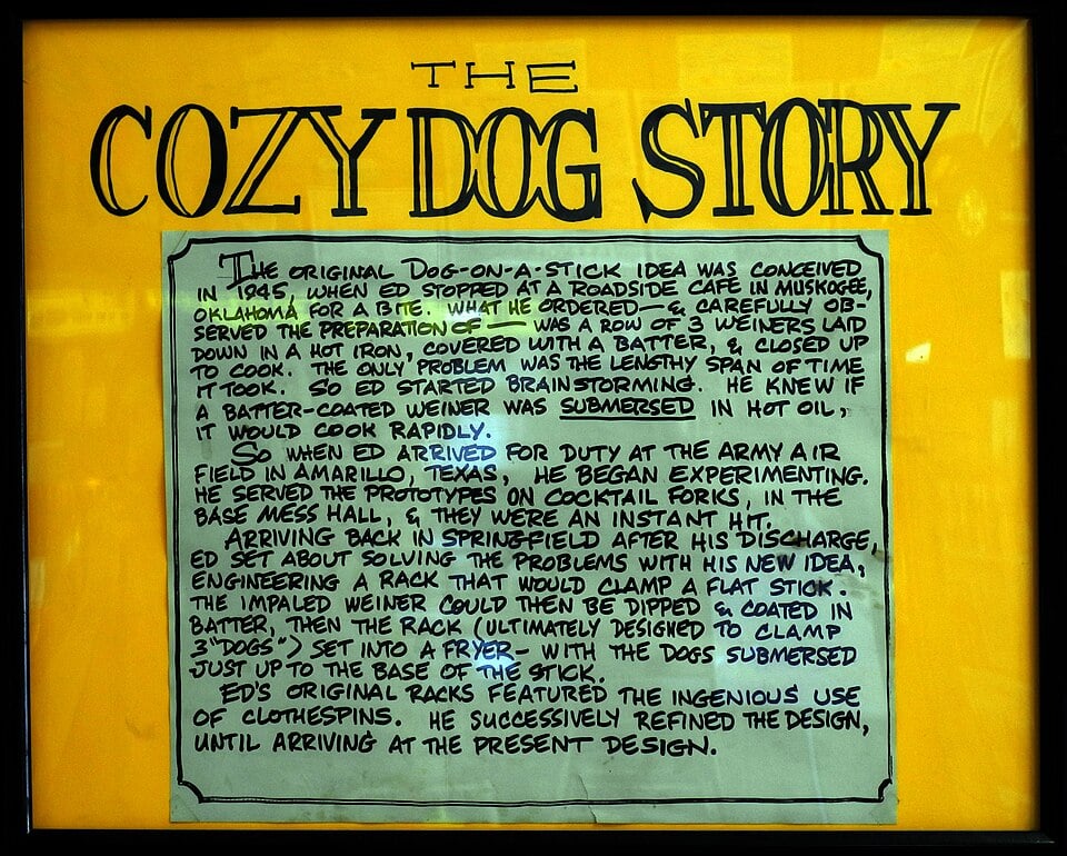 A framed sign titled "The Cozy Dog Story" describes how Ed Waldmire Sr. invented the Cozy Dog, a hot dog dipped in cornmeal batter and deep-fried, inspired by his army experience. The sign has handwritten text on a yellow background.