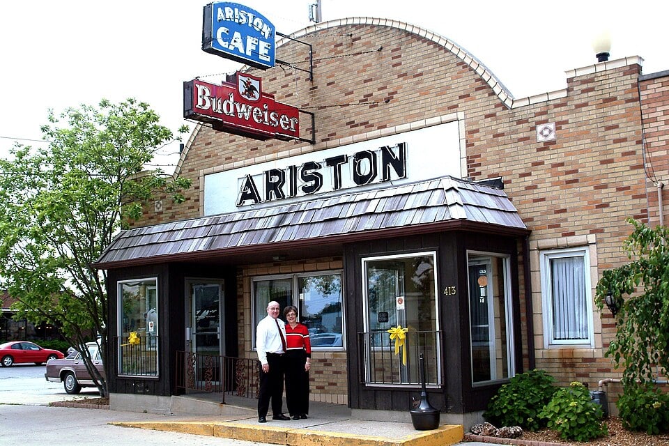 Two people stand smiling in front of the Ariston Café, a historic brick restaurant with large windows and vintage signs for Ariston and Budweiser above the entrance. Trees and parked cars are visible nearby.
