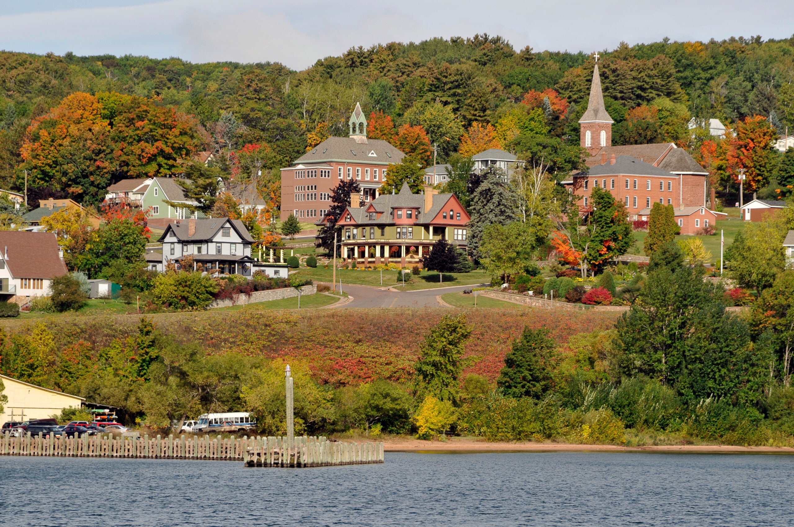 A scenic view of a small town with historic buildings, houses, and a church steeple amid colorful autumn trees, overlooking a waterfront with a dock and calm water in the foreground.