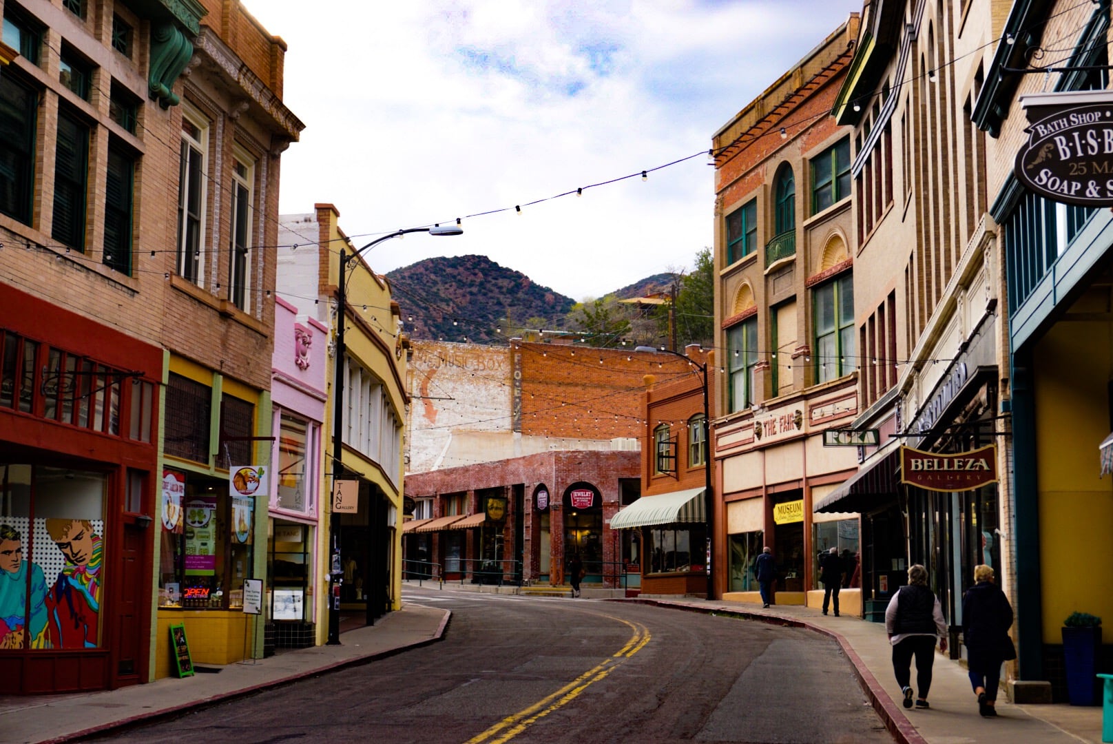 A colorful, slightly curved street lined with historic buildings and shops in a small town, with two people walking on the sidewalk and hills visible in the background under a partly cloudy sky.