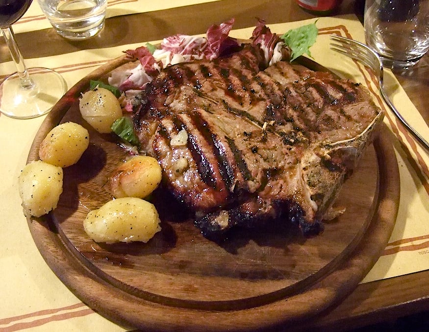 A grilled T-bone steak served on a wooden platter with a side of roasted potatoes and fresh salad greens, accompanied by a fork and a glass of water in the background.