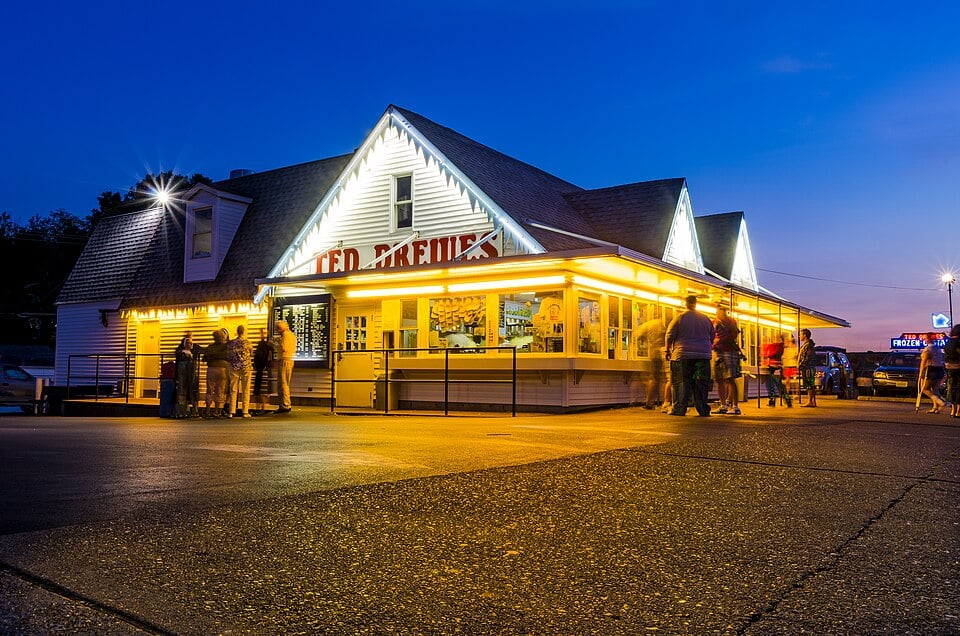 A brightly lit ice cream stand at dusk, with people gathered outside under the glowing lights. The sky is deep blue, and the building’s sign reads “Ted Drewes.” The scene gives a warm, inviting atmosphere.