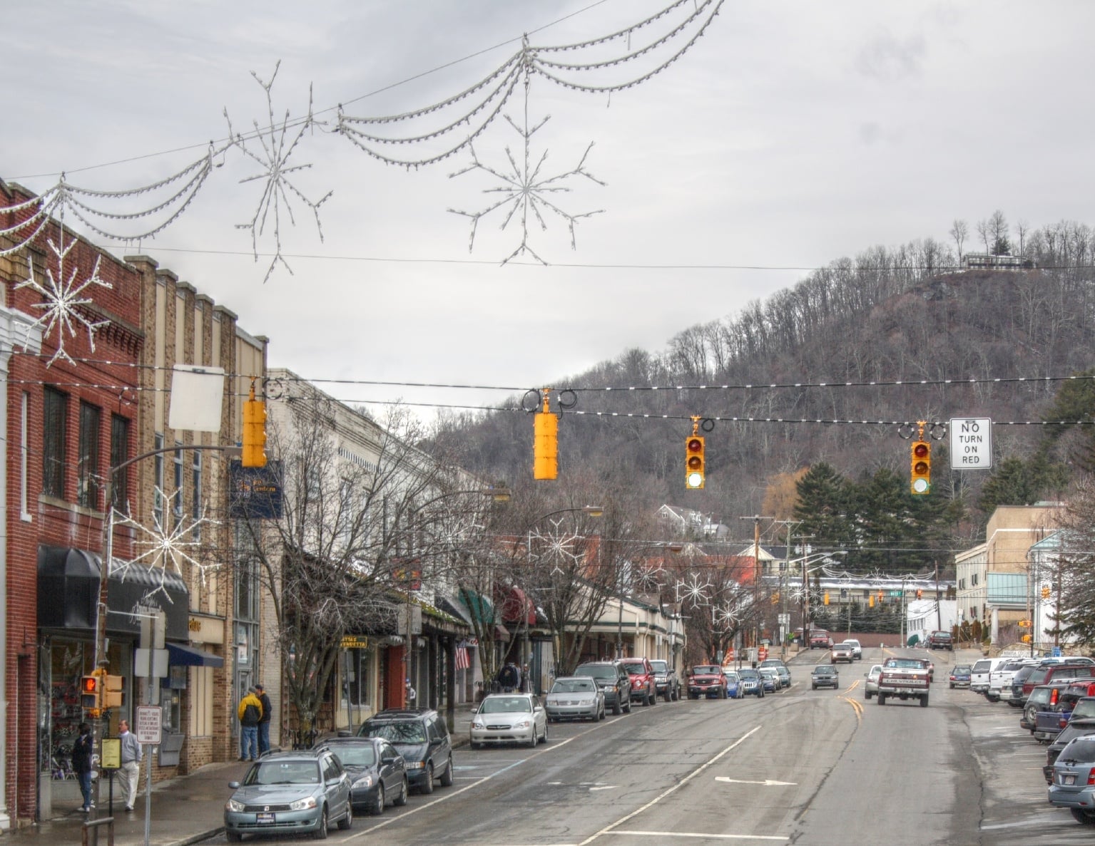 A small town street lined with shops and parked cars, decorated with large hanging snowflakes and string lights. Traffic lights are red, and tree-covered hills are visible in the background under a cloudy sky.