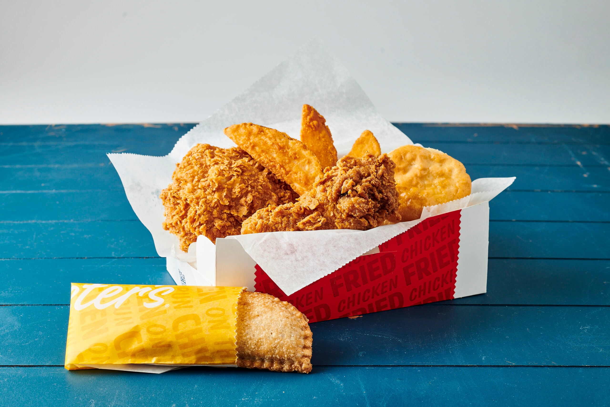 A meal box from popular fried chicken chains with crispy chicken pieces, potato wedges, and a biscuit sits on a blue wooden table. In front, there is a packaged fried pie in a yellow wrapper.