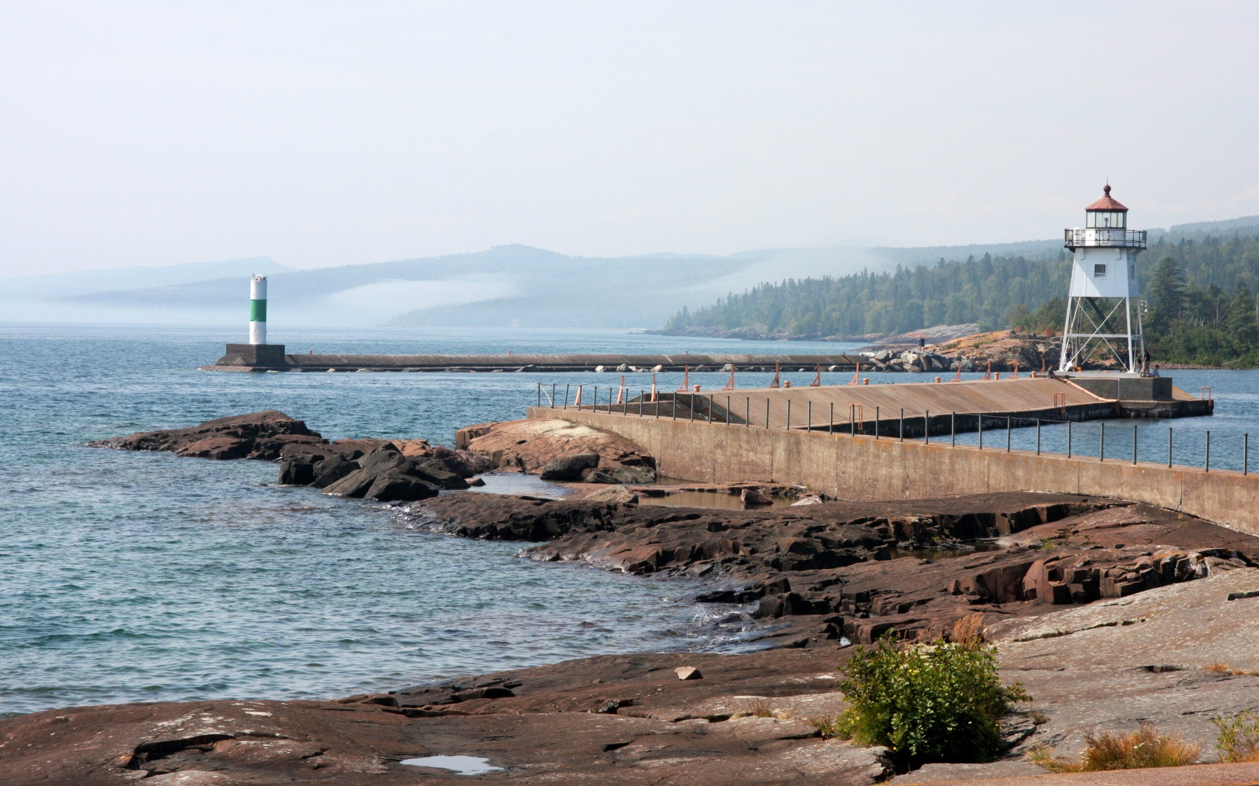 A long pier extends into a blue lake, ending at a white lighthouse with a red roof. Another small green-and-white beacon stands at the far end, with distant forested hills in the background under a hazy sky.